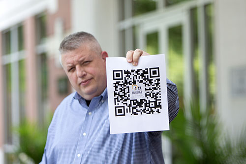 New Posters, QR Code to Help Find Missing Kids Image of John Bischoff II standing outside NCMEC withshort cropped hair and a blue checked button down shirt. He hold out a paper toward the viewer with a QR code and the NCMEC logo.